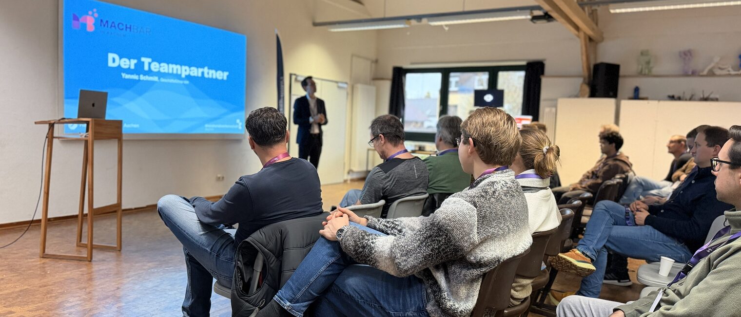 Eine Gruppe von Menschen sitzen vor einem Redner. Im Hintergrund sieht man eine Präsentation mir der Folie "Der Teampartner" 
